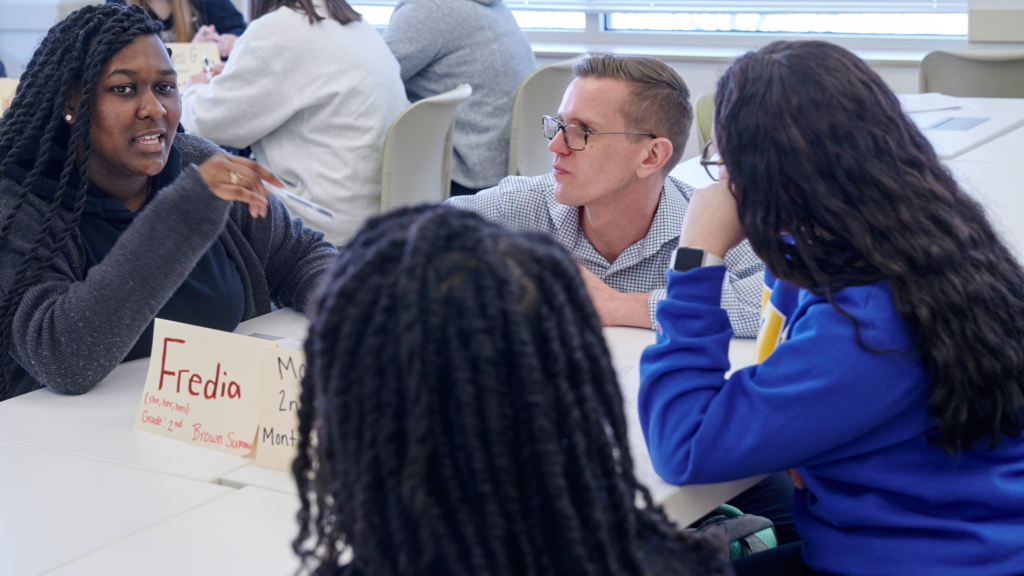 Instructor Ryan Hughes engages a group of his students in conversation around a table in class