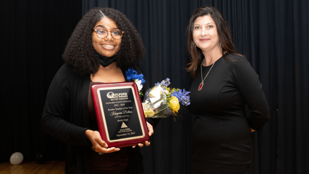 Guilford County Schools Rookie Teacher of the Year Kenyona Bethea holds her plaque and flowers while standing with GCS Superintendent Whitney Oakley in front of a black background