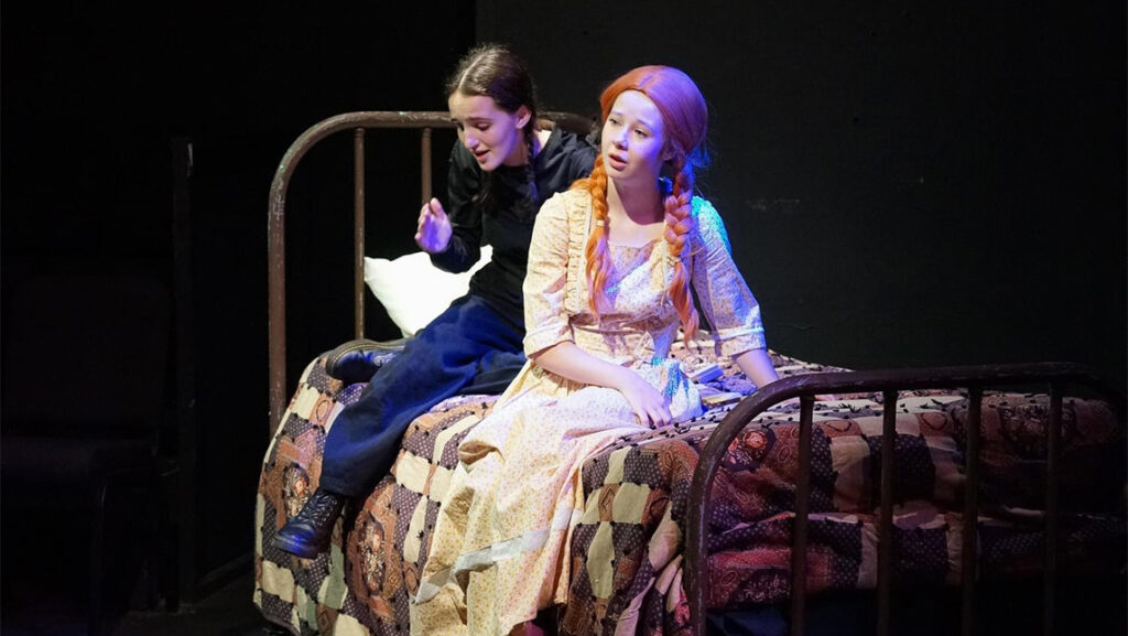 An actress playing Anne of Green Gables sits on a bed while a UNCG student in black clothes shadows her performance