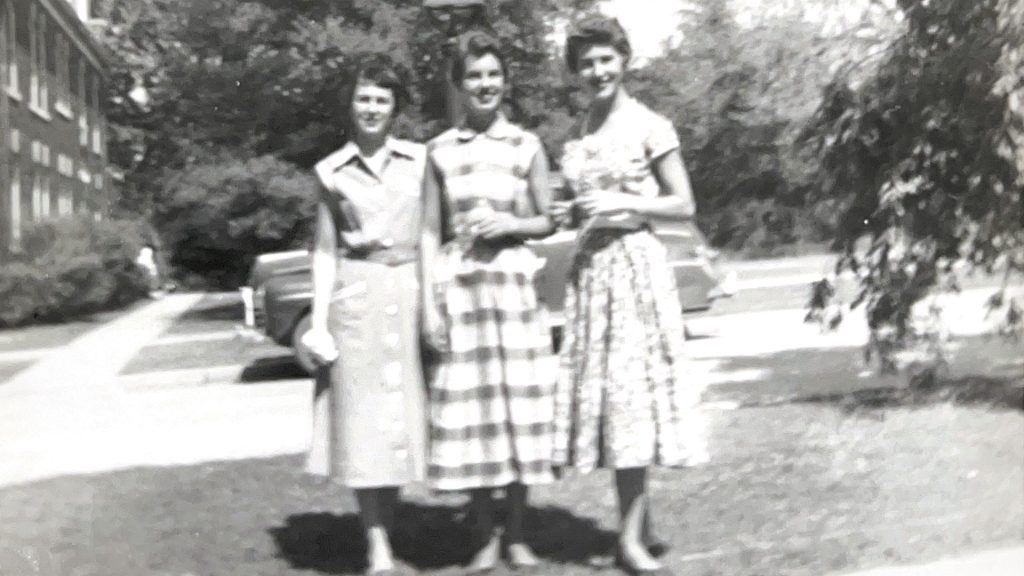 Black and white photo of three young women standing outdoors in front of a parked car and trees. They are dressed in mid-20th-century style dresses. The woman in the middle, Barbara Berryhill, is wearing a checkered dress, while the others wear patterned or solid dresses. The background includes a sidewalk, a brick building on the left, and leafy trees on the right. The image has a vintage, slightly blurry quality.
