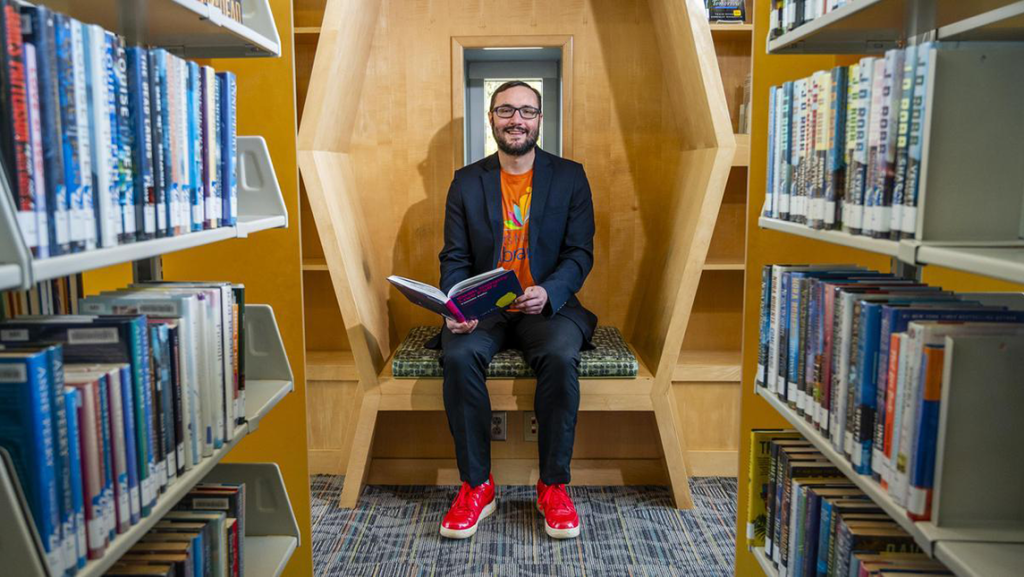 Dr. Noah Lenstra sits in a uniquely shaped wooden reading nook between two bookshelves in a library. He is smiling, holding an open book, and wearing a dark suit, a bright orange t-shirt with a colorful logo, and red sneakers. The scene is well-lit and framed by rows of books on either side.