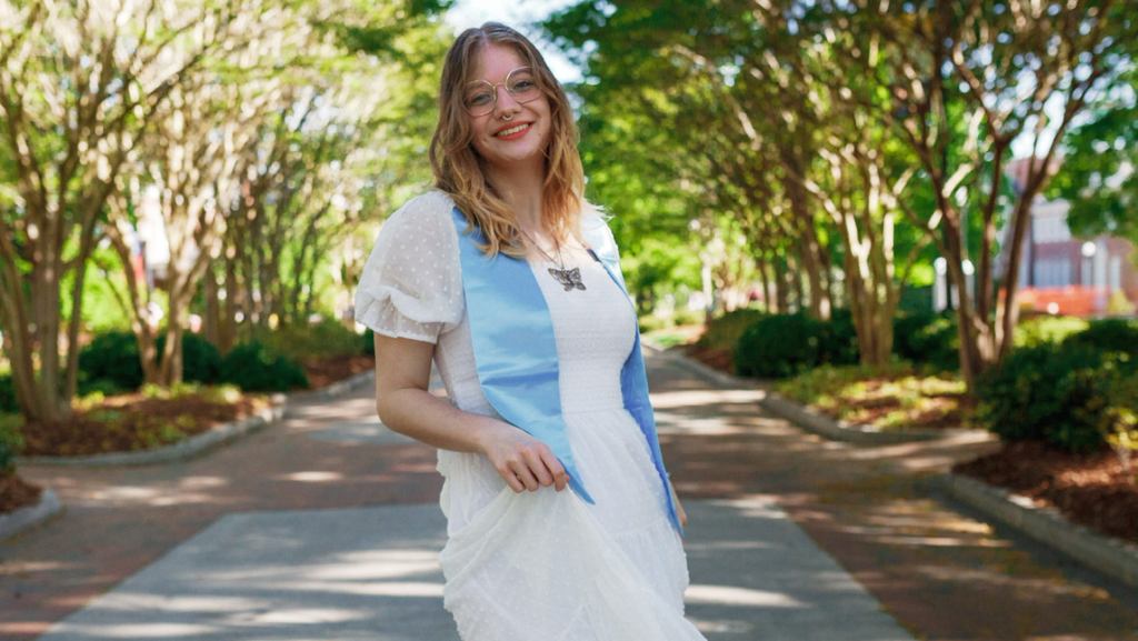 Alyssa Causey, wearing a white dress and blue stole, smiles on a sunny, tree-lined path.