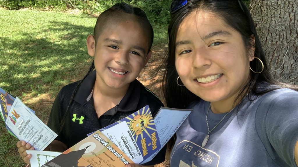 Maria Rodriguez Campos and a young boy smile while sitting outside in the grass. The boy holds several colorful certificates, and the woman leans in close for the photo.