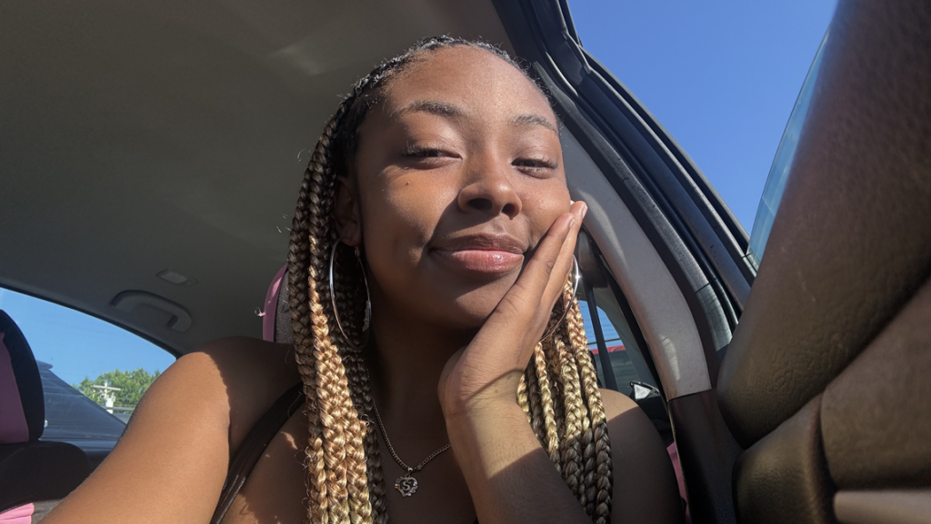 Sadaria Lewis sitting in a car, smiling softly with her hand resting on her cheek, sunlight shining on her face.