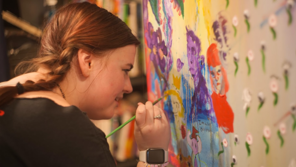 Ava Cobb, with braided hair, paints a colorful mural featuring abstract and cartoon-like faces in bright colors. She is holding a paintbrush and concentrating on her work.