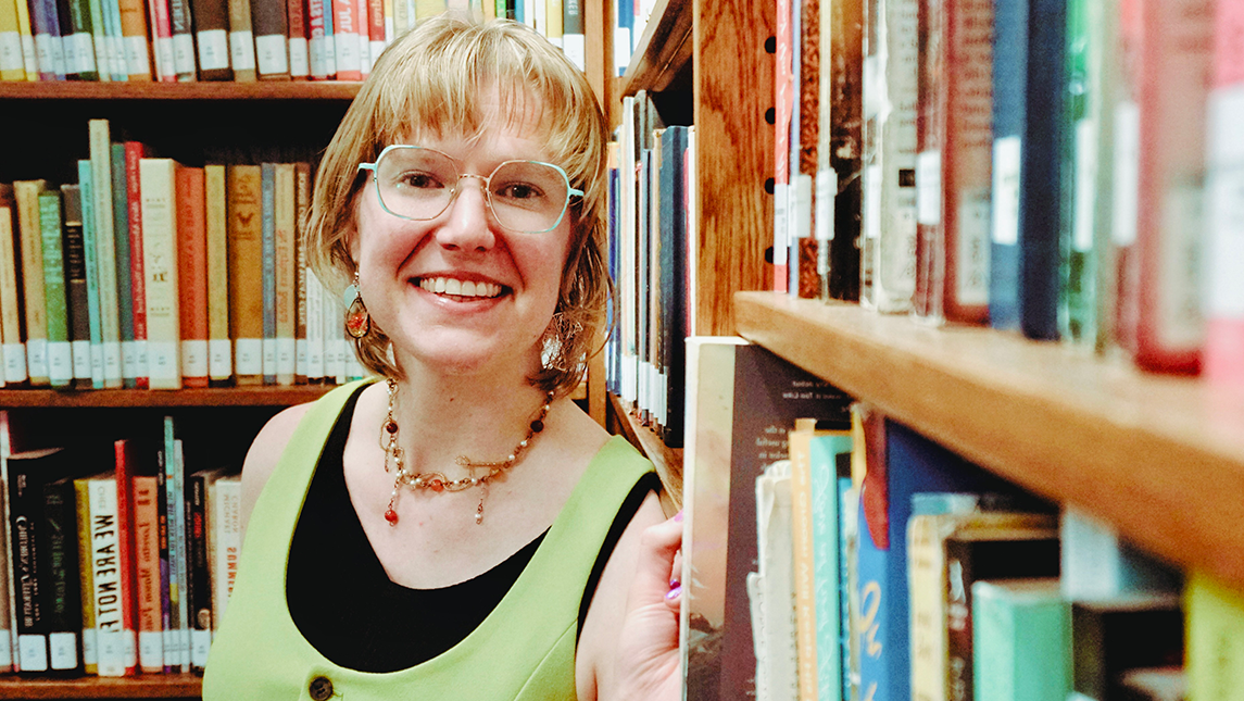 H. Blake-Lee, with short blond hair and light blue glasses, stands in a library aisle, holding a book on a shelf. She is wearing a light green top over a black shirt, decorative earrings, and a beaded necklace. Rows of colorful books fill the shelves around them.