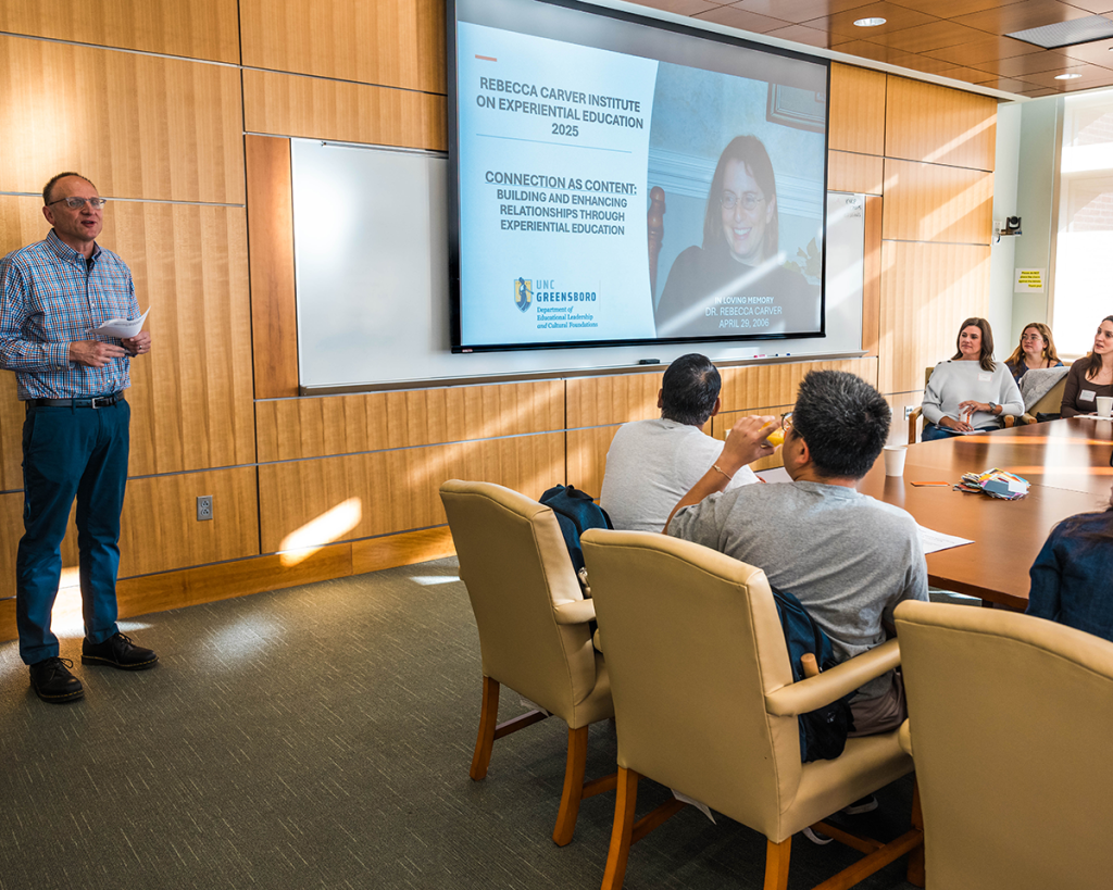 A conference room with a large wooden table surrounded by beige chairs, where several people are seated and facing a wall-mounted screen. The screen displays a presentation titled “Rebecca Carver Institute on Experiential Education 2025” with the subtitle “Connection as Content: Building and Enhancing Relationships Through Experiential Education” and the UNC Greensboro logo. A person stands to the left of the screen holding papers, while natural light streams in from windows on the right side of the room.