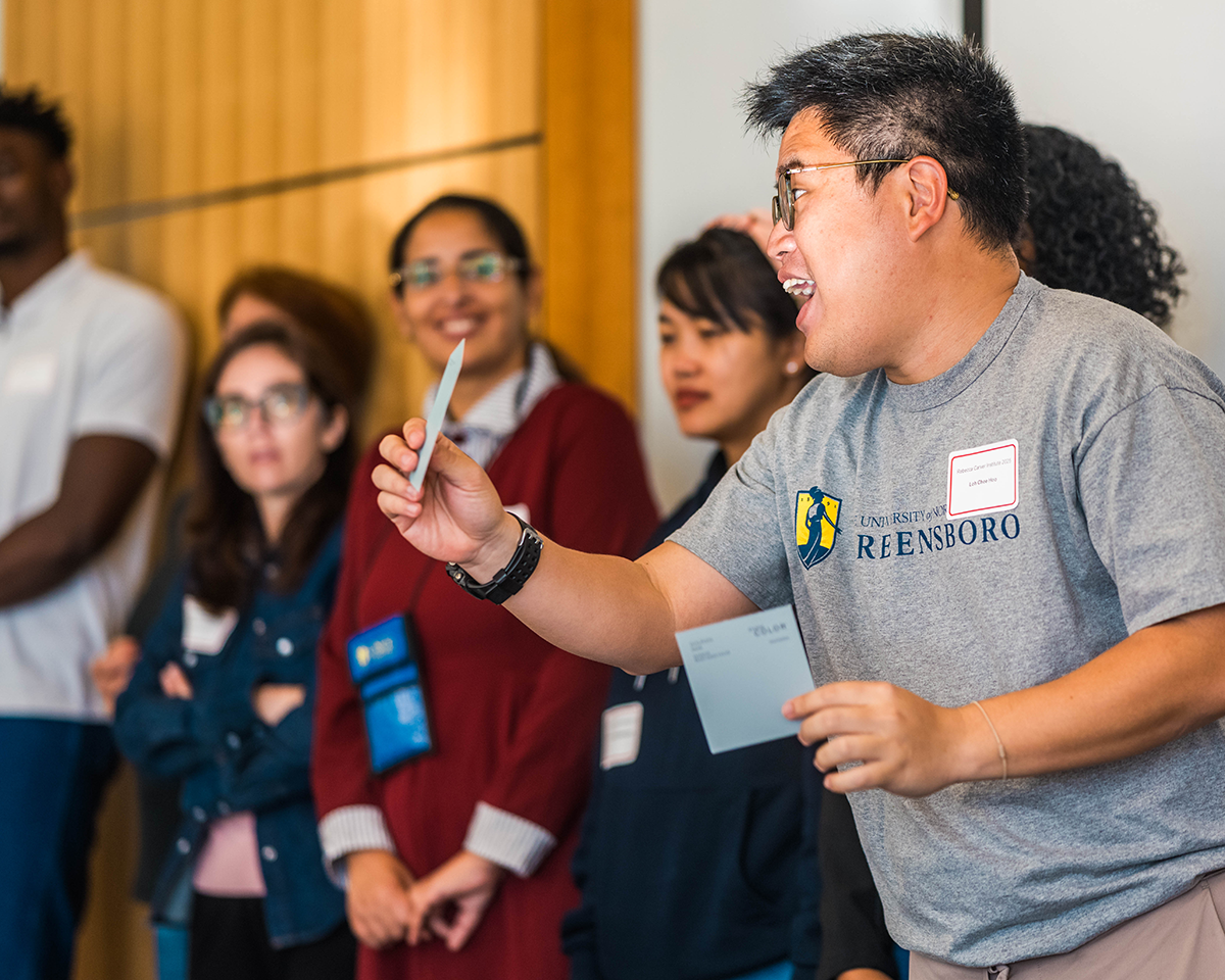 A person wearing a UNC Greensboro T-shirt enthusiastically holds up a card while speaking during an interactive workshop. Several other participants stand in the background, watching and smiling in a classroom setting.