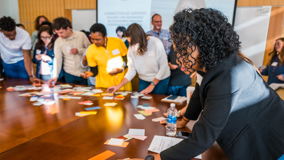 A diverse group of Rebecca Carver Institute attendees gather around a large table covered with colorful paper cards, engaging in a collaborative activity during a workshop. One participant in the foreground leans forward, observing the discussion closely, while others reach for cards and interact with one another in a bright, modern classroom setting.