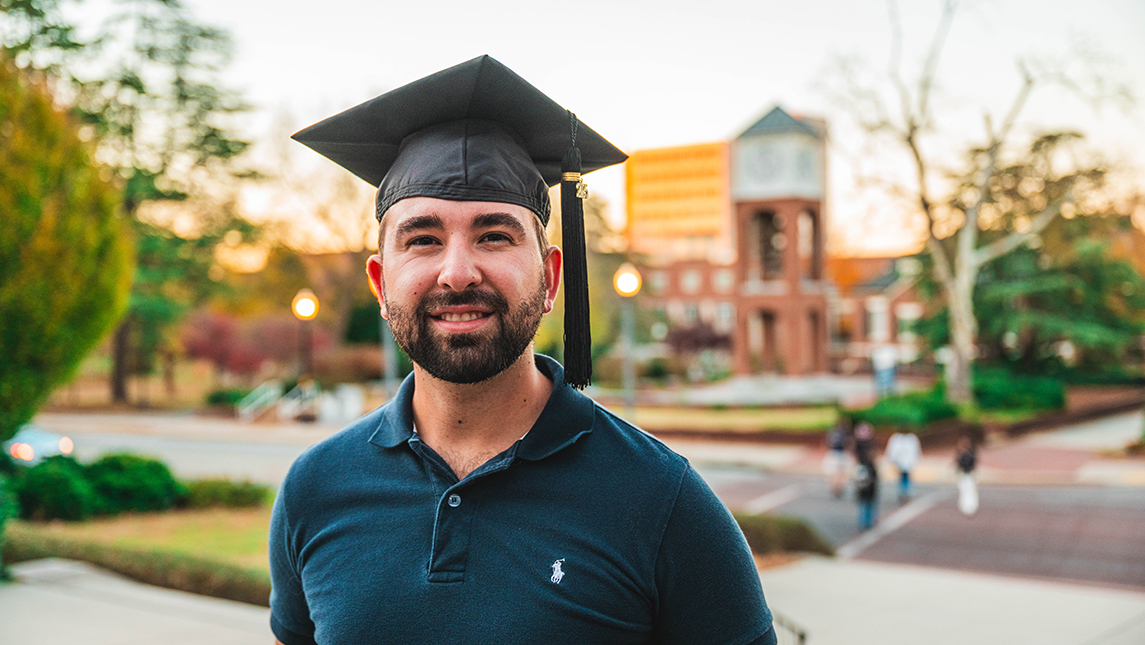 Caleb Kirk stands in a graduation cap on UNCG's campus