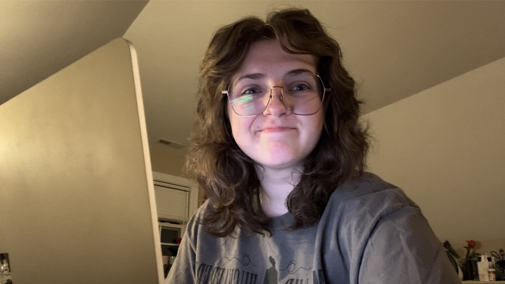 Katelyn Cruthirds, with shoulder-length wavy hair and round glasses, smiles at the camera while sitting indoors at a desk, lit by warm light against a neutral background