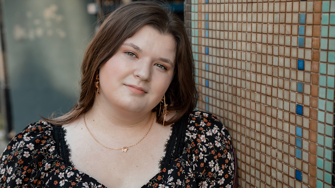 Skylar Sumrell leans against a tiled wall, looking at the camera with a calm expression. They are wearing a black floral-patterned top, gold earrings, and a delicate necklace. The background features small square tiles in neutral and blue tones.