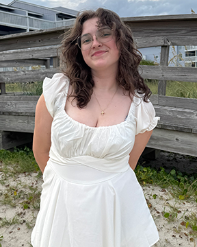 Katelyn Cruthirds, wearing a white short-sleeved dress stands outdoors on sandy ground, smiling slightly, with a wooden boardwalk and grassy dunes behind them under a cloudy sky.