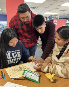 A teacher and three students lean over a table, reading and pointing to a book together during a small-group learning activity in a library.
