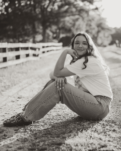 Black-and-white photo of Abigail Hartley sitting on a gravel path beside a wooden fence, smiling at the camera. She rests her chin on her hand and wears a T-shirt, jeans, and sandals, with trees softly blurred in the background.
