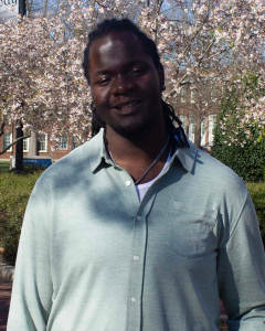 Grady Dupree-Isaac wears a light-colored shirt while standing in front of flowering trees