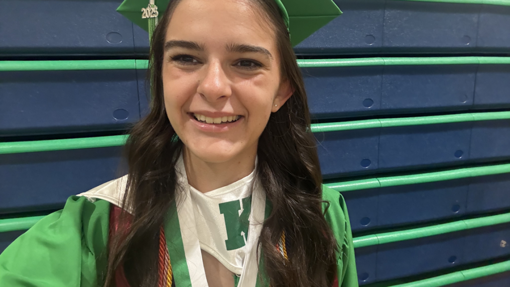 A smiling Megan Fongemy wearing a green cap and gown with a 2025 tassel and a white stole featuring a green “K,” standing in front of blue and green gym bleachers.
