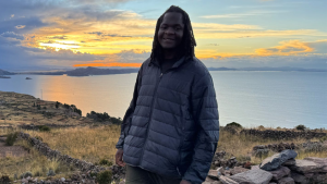 Grady Dupree-Isaac wears a dark jacket and stands on a hillside overlooking a large body of water at sunset, with golden light reflecting off the surface and distant mountains silhouetted against a colorful sky.