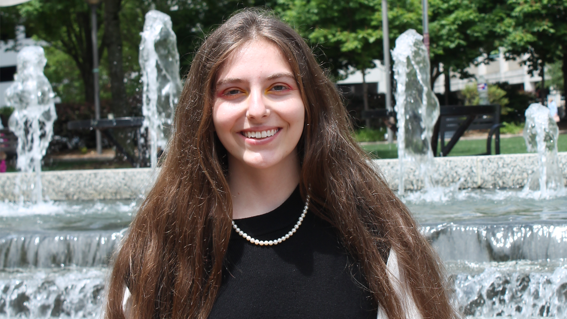 Leila Lovell, with long brown hair and wearing a black top and a pearl necklace, standing in front of a tiered outdoor fountain with water spraying upward and trees in the background.