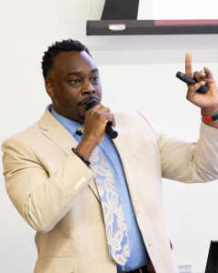 Dr. James E. Ford, wearing a beige suit and patterned tie, speaks into a handheld microphone while raising one finger, holding a presentation remote, in a bright indoor setting.