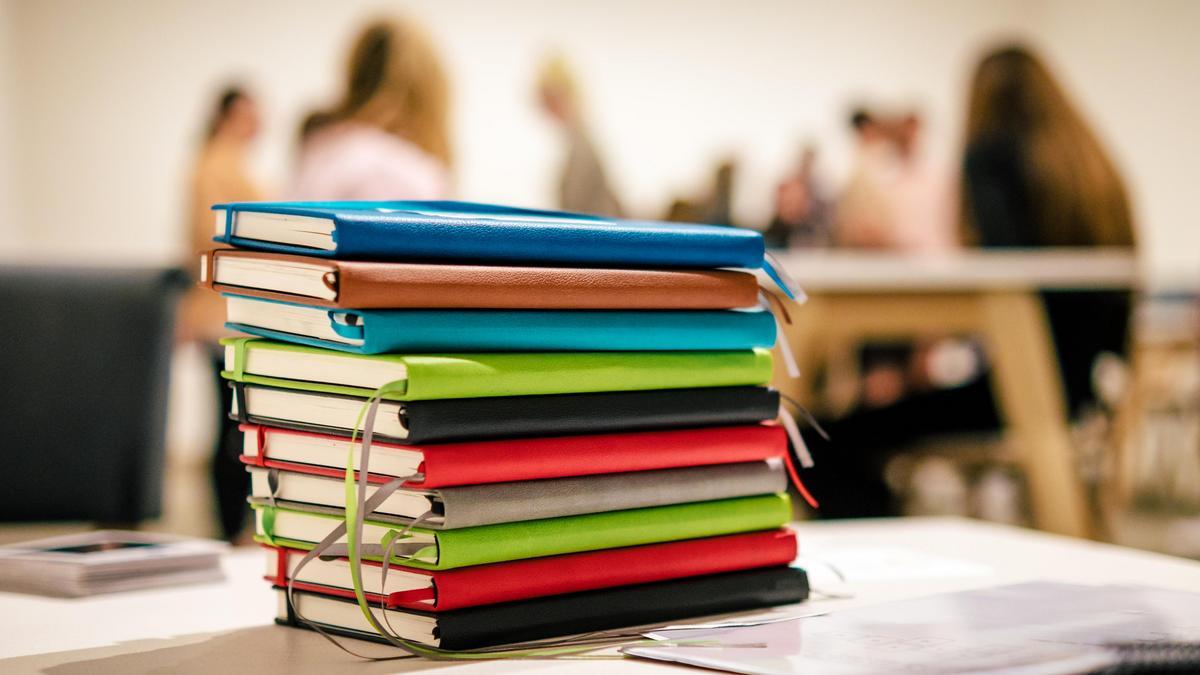 Stack of different colored books sits on a table with students in the bacground