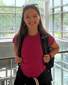 Sawyer Wemyss stands in the open lobby area of the School of Education Building, wearing a bright pink T‑shirt, black pants, and a backpack, with long hair and glasses resting on the head, facing the camera