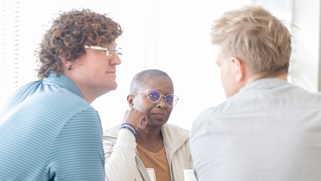 Three adults sit together in conversation at a table during Brock's Critical Conversations Conference; a woman with short gray hair and glasses looks attentively toward a man speaking, while another person listens nearby in a bright indoor setting.