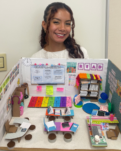 Yana Gaitan stands behind her detailed miniature classroom model displayed on a table. The model includes small desks, books, a reading corner, a colorful alphabet and number rug, and a “Good Morning” board with classroom schedules and notes. Decorations like posters, sticky notes, and a “READ” sign line the walls, while tiny props such as laptops, notebooks, and seating areas create a realistic classroom layout.