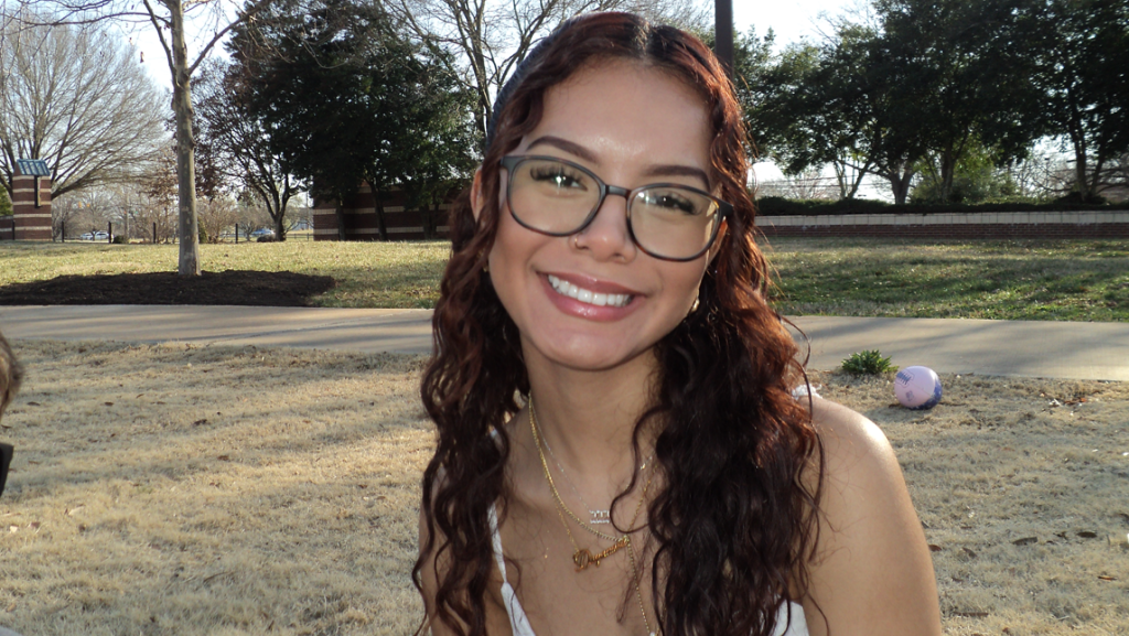 Yana Gaitan with long wavy brown hair and glasses looks at the camera while sitting outdoors on a sunny day. She wears a white top and gold necklaces. Behind her are trees, a sidewalk, and a grassy area, with a small pink ball visible in the background.