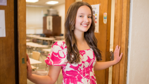 A smiling Sawyer Wemyss in a pink-and-white floral dress stands in a classroom doorway, holding the door open with both hands, with desks and chairs visible behind her.