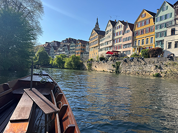 Wooden boat in the foreground on a calm river, with a row of colorful, historic half-timbered houses lining the right bank. The buildings feature pastel facades of yellow, peach, and cream, with steep roofs and many windows, set above a stone embankment. Trees and lush greenery fill the left side of the scene, partially shading the water. The sky is clear and bright blue, and strong sunlight reflects off the rippling water, suggesting a peaceful daytime setting in a scenic European riverside town.