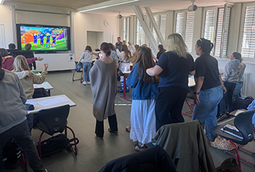 Students and instructors stand and move around a modern classroom during an interactive group activity. A colorful educational soccer game is displayed on a large screen at the front of the room while participants engage with one another beside desks and chairs.