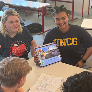 Group of students seated around a table in a classroom, with two individuals facing the camera. One person on the right wears a navy shirt with large yellow “UNCG” lettering and holds a tablet displaying an image. Another person on the left wears a dark T-shirt with a circular red-and-white graphic. In the foreground, two additional students are partially visible from behind, looking toward the tablet and a printed worksheet on the table covered with handwritten notes. The classroom includes tables, chairs, and personal items like a backpack, with bright indoor lighting and a casual, collaborative setting.