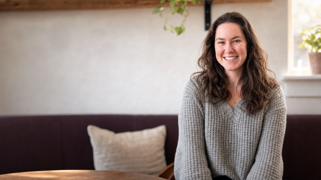 Portrait of a smiling woman with long wavy brown hair, seated on a bench indoors, wearing a gray knit sweater.
