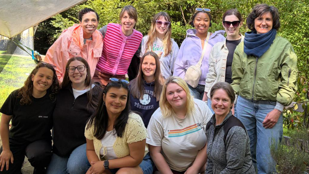 Group photo of UNCG education exchange participants and faculty members posing outdoors in a garden setting during a study abroad trip in Germany. The group smiles at the camera while standing and kneeling together near greenery and a canopy-covered seating area.