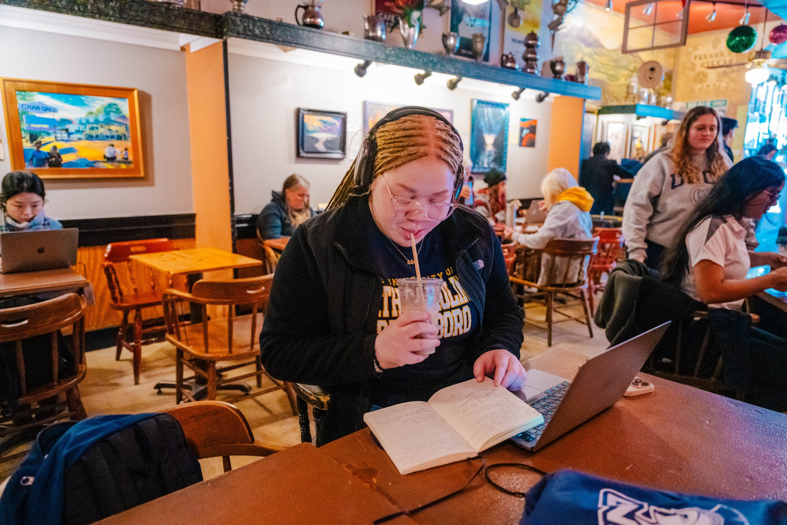 Person wearing headphones sits at a wooden table in a busy café, drinking from a cup while reading notes beside an open laptop; colorful artwork and other patrons are visible in the warmly lit background.