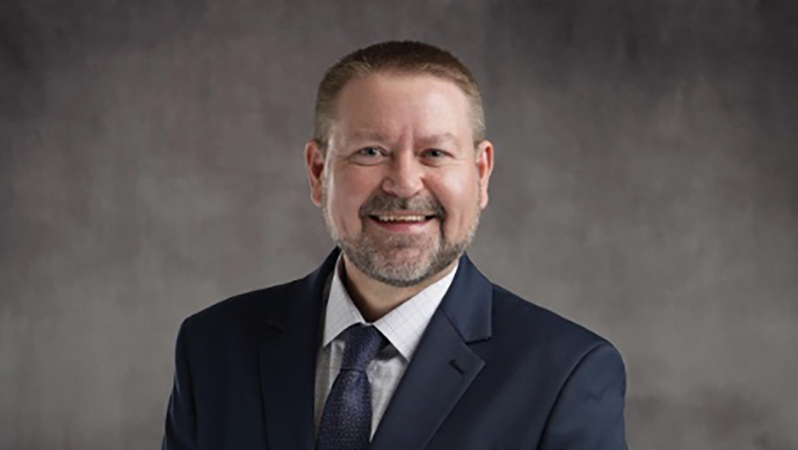 Dr. Charles Perkins wearing a suit and tie in front of a gray background