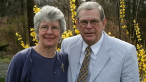 Tommy and Nancy Teague stand together outdoors, smiling at the camera. Nancy wears a navy top and cardigan, while Tommy wears a light-colored blazer, dress shirt, and patterned tie. Yellow flowering shrubs and trees in the background create a springtime setting.