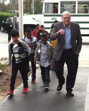 Tommy Teague, in a grey blazer and spectacles, smiles as he walks alongside a group of young school-aged children on a paved path, with a white bus visible in the background.