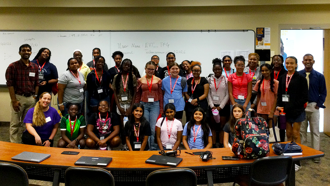 A group of approximately 30 middle school students and several camp leaders pose together in a university classroom during the Find Your STEM, Shape Your Future camp. The students, wearing red lanyards and name badges, stand and kneel in front of a whiteboard while laptops and backpacks sit on tables in the foreground. The group smiles for a commemorative photo celebrating their participation in the STEM program.