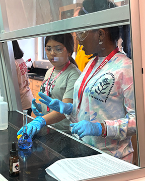Two middle school students wearing safety goggles, gloves, and camp lanyards conduct a science experiment at a laboratory workstation. One student carefully holds a flask while the other observes and gestures as they follow a printed lab guide.