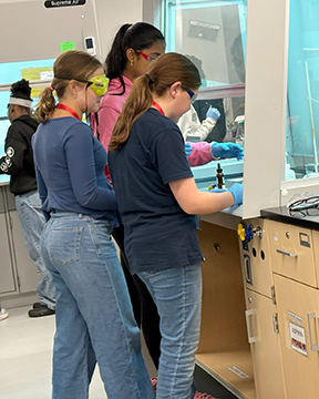Three middle school students wearing safety goggles and gloves work together at a laboratory bench during a hands-on STEM activity. The students focus on their experiment inside a fume hood while following lab procedures.