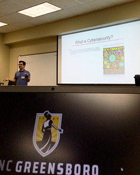 A presenter delivers a lesson on cybersecurity to students in a university classroom. A presentation titled "What is Cybersecurity?" is projected on a screen, while a UNCG-branded computer monitor is visible in the foreground.