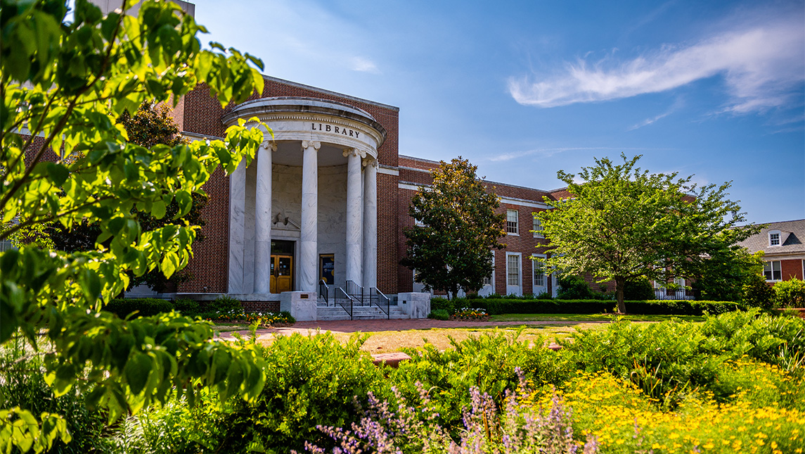 Front view of a brick campus library with white columns, framed by leafy trees and landscaped gardens on a sunny day.
