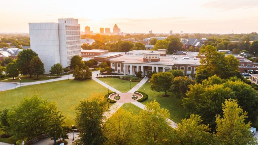 Aerial view of the UNCG campus at sunset, with academic buildings, green lawns, tree-lined walkways, and a city skyline in the background.