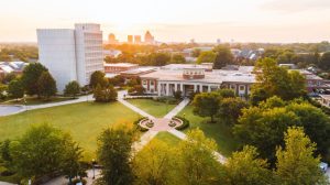 Aerial view of the UNCG campus at sunset, with academic buildings, green lawns, tree-lined walkways, and a city skyline in the background.