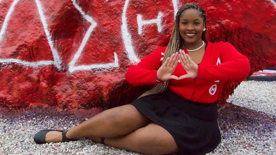 Syneece Forbes in a red Delta Sigma Theta sweater, seated before a red painted rock and forming a triangle with her hands.