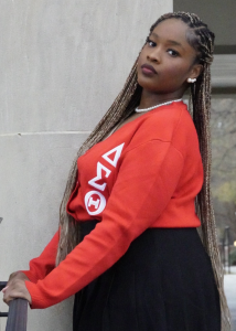 Syneece Forbes in a red Delta Sigma Theta sweater, standing by a railing with long braids and pearl jewelry.
