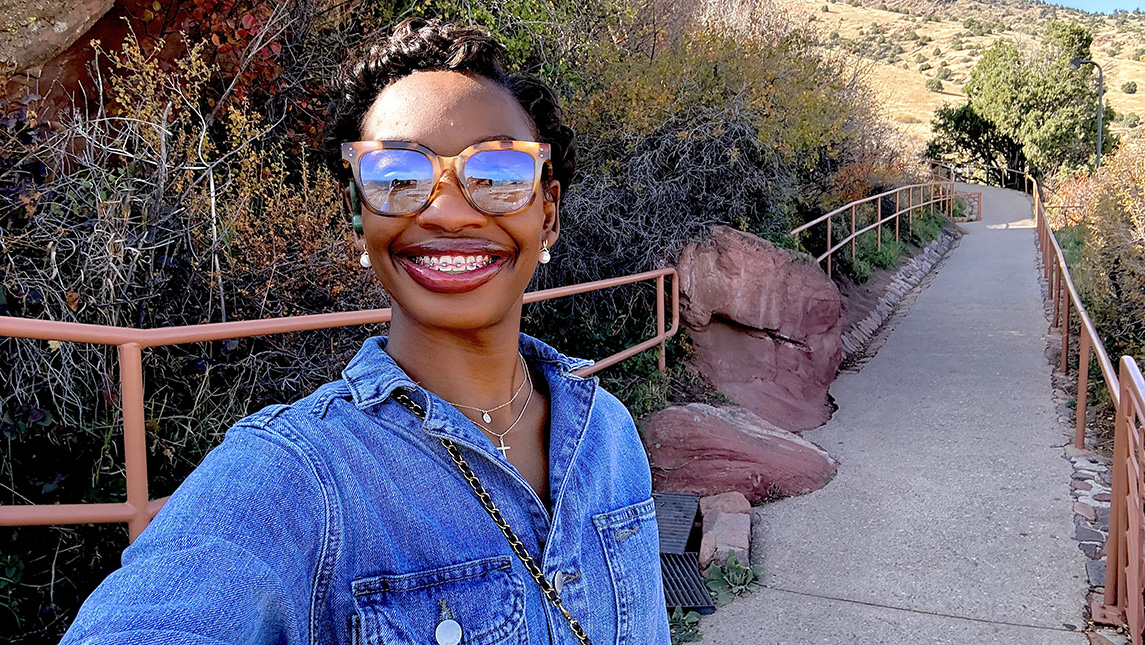 Adrianna Fields takes a smiling selfie on a hillside path bordered by red rocks and railings.