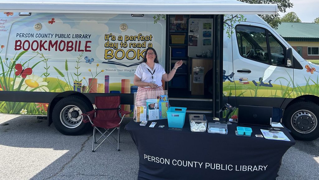 Dawn Gaug, a Person County Public Library staff member, stands beside a book display and outreach table in front of the colorful library bookmobile.